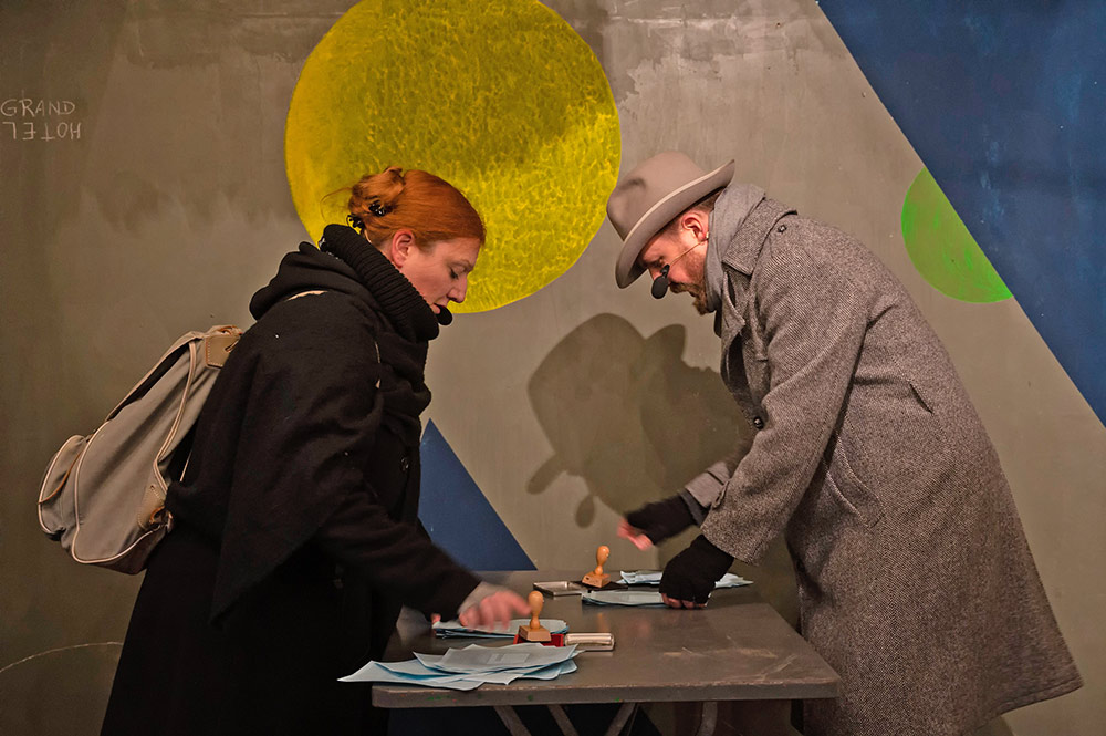 Im Bild sind zwei Schauspieler*innen mit Rucksack und Hut. Zwischen ihnen ist ein Tisch mit vielen blauen Papieren. Sie sind dabei diese zu bestempeln. Im Hintergrund ist eine Wand mit geometrischen Formen in gelb und blau.