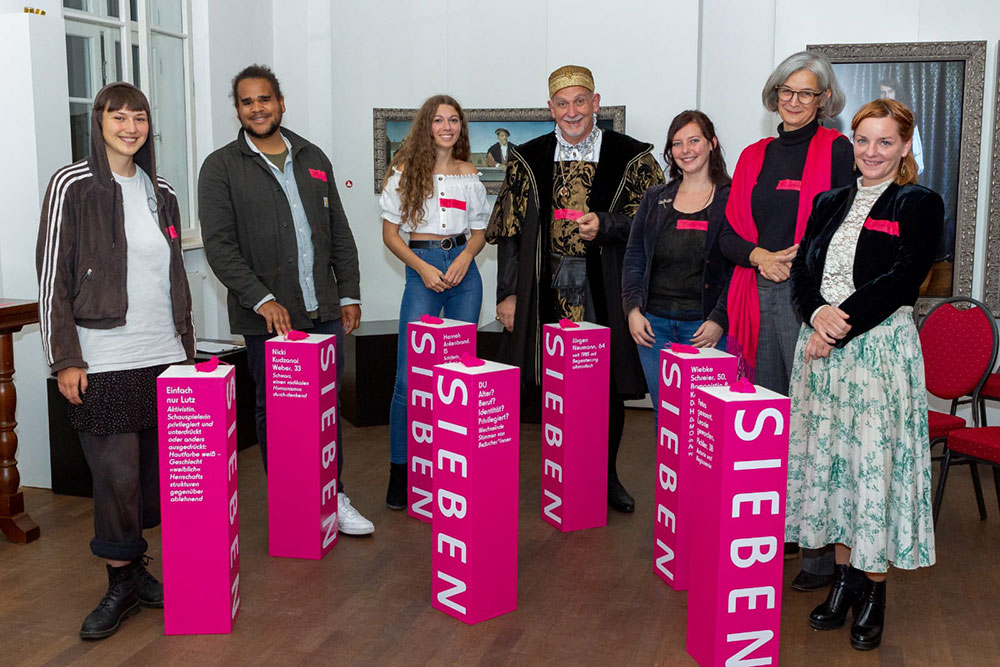 Im Foto sind sieben Menschen die hinter sieben Magenta-Säulen mit der Aufschrift SIEBEN stehen.