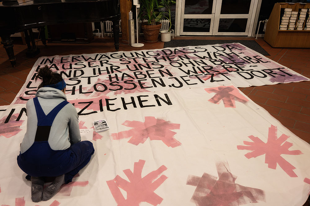 Das Foto zeigt zwei große Plakate, die gerade beschrieben werden. Darauf steht bereits "IN ERWÄGUNG DASS DA HÄUSER STEHEN WÄHREND IHR UNS OHNE BLEIBE LASST HABEN WIR BESCHLOSSEN JETZT DORT EINZUZIEHEN"