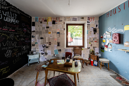 Auf dem Foto ist ein Raum mit einem Tisch in der Mitte zu sehen. Die jeweils schwarze und blaue Wand links und rechts sind bunt beschrieben. Die weiße Wand in der Mitte ist beklebt mit vielen ausgedruckten Zetteln.