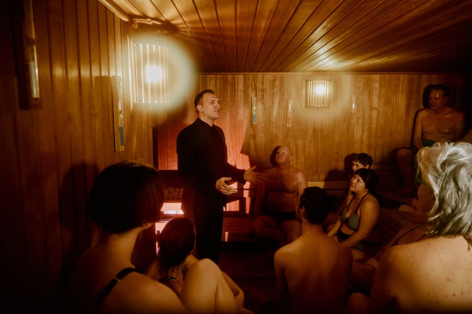 Das Foto entstand in einer Sauna. Im Publikum steht ein schwarz gekleideter Schauspieler, der eine Rede hält.