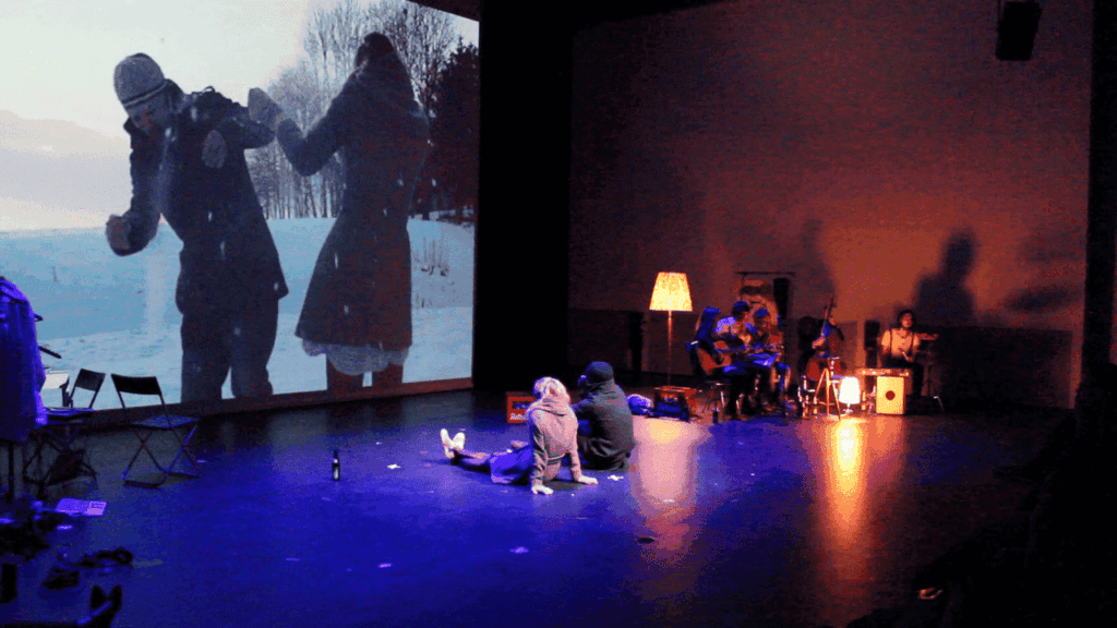 Auf einer Bühne sitzen zwei Menschen vor einer Wandprojektion, die Menschen im Winter zeigt. Sie sind in einem dunklen Raum und im Hintergrund sitzt eine Band.