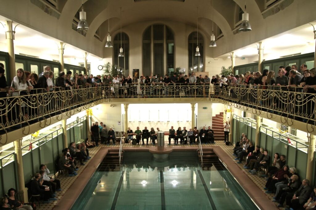Auf dem Foto ist ein großes Stadtbad zu sehen. Viele Menschen sitzen um das Becken oder stehen oben am Geländer einer Galerie.