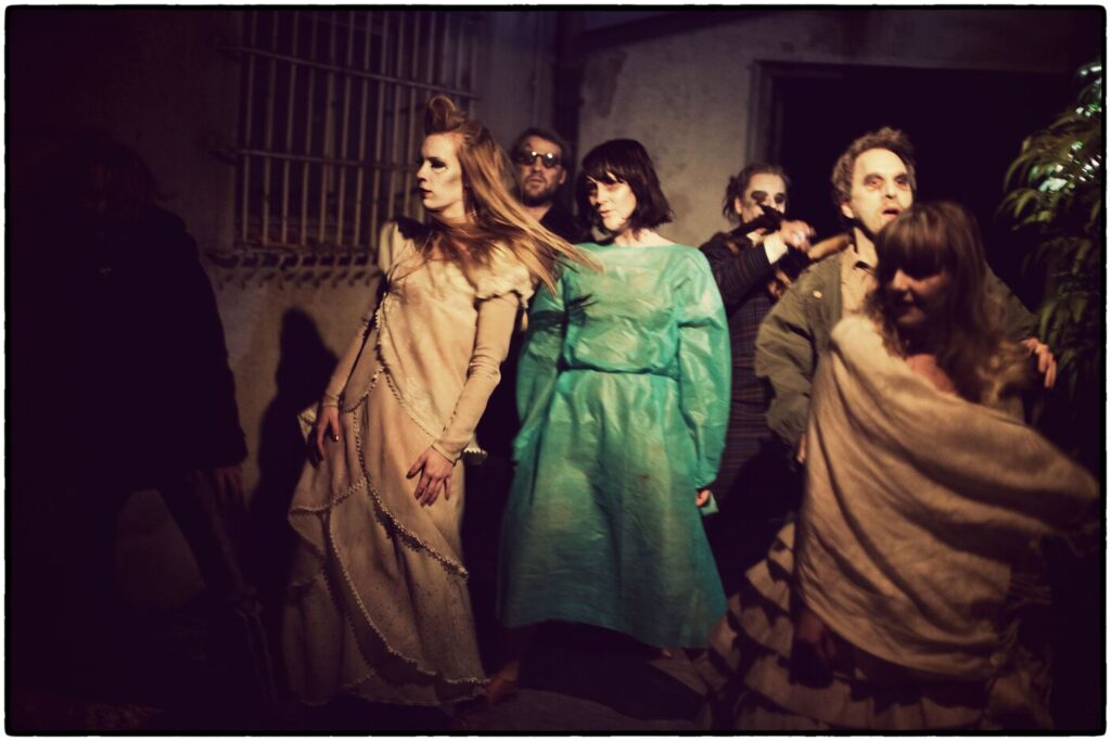 Auf dem Foto sind mehrere verkleidete, weiß geschminkte Menschen in langen Kleidern. Sie sind in einem dunklen Raum.
