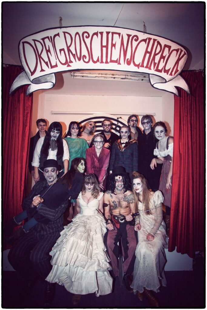 Auf dem Gruppenfoto sind mehrere kostümierte Menschen zwischen roten Vorhängen in weißen Kleidern, schwarzen Anzügen und roten Outfits. Sie sind weiß geschminkt und über ihnen hängt eine schwungvolle Schrift in rot "DREIGROSCHENSCHRECK".