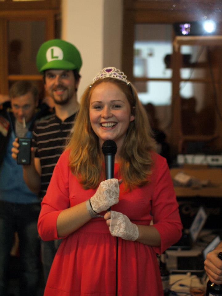 Auf dem Foto ist eine glückliche Frau in pinken Kleid und Krone mit Mikrofon. Hinter ihr steht ein Mann mit "Luigi-Kappe".