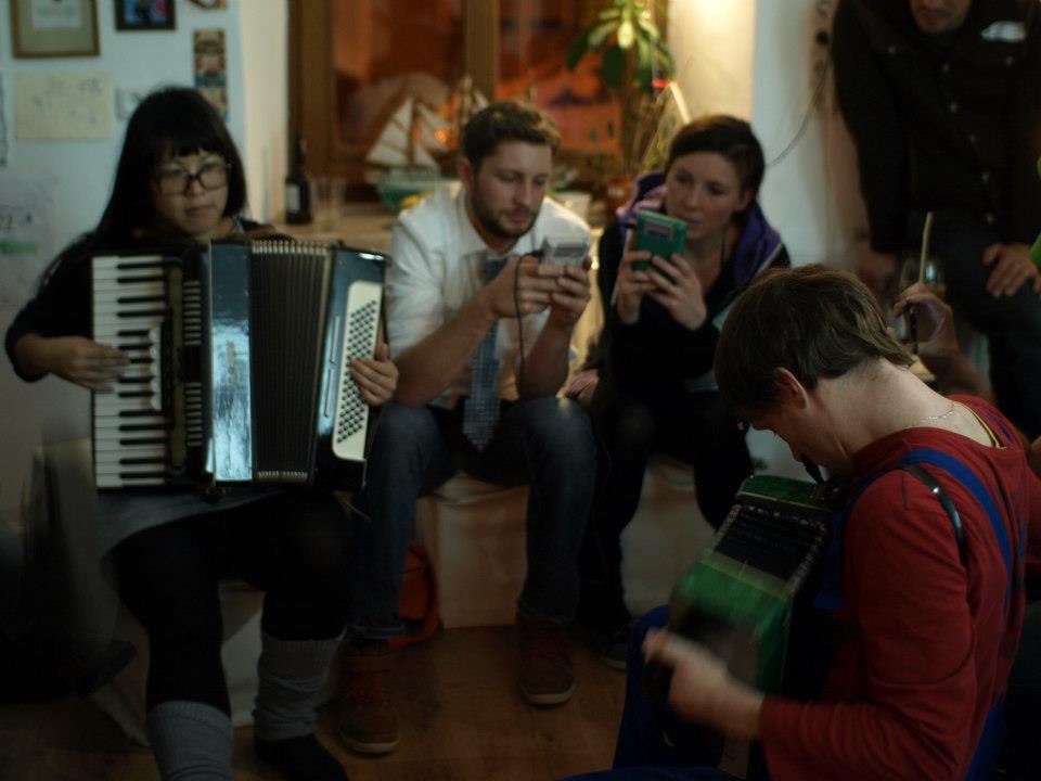 Auf dem Foto sitzen zwei Menschen, die an Gameboys spielen. Links und rechts von ihnen sitzen zwei, die Ziehharmonika spielen.