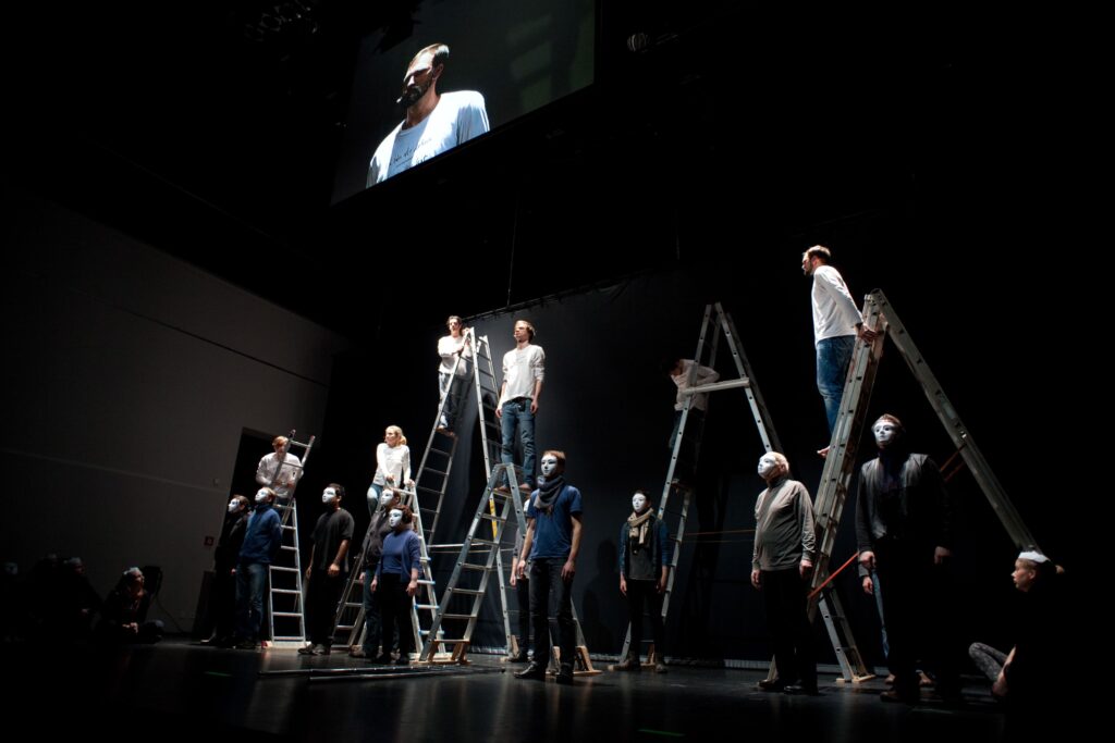 Auf dem Foto sind mehrere Menschen mit weißen und blauen Oberteilen und Masken, die auf oder um Klappleitern stehen. Der Raum ist abgedunkelt.