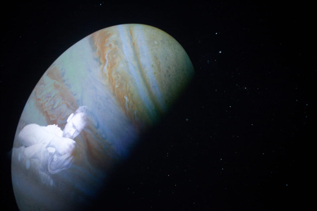 Auf dem Bild sieht man einen Sternenhimmel mit einem Planeten auf dem die Reflektion eines Mannes zu erkennen ist.