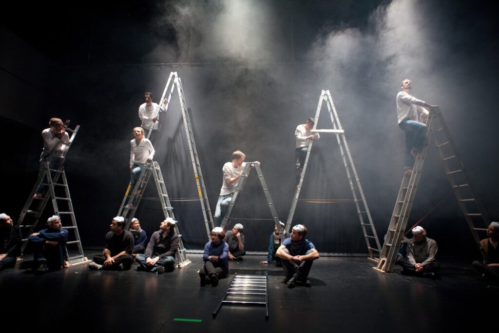Auf dem Foto sind mehrere Menschen mit weißen und blauen Oberteilen, die auf oder um Klappleitern stehen oder sitzen. Der Raum ist abgedunkelt und Nebel in der Luft.