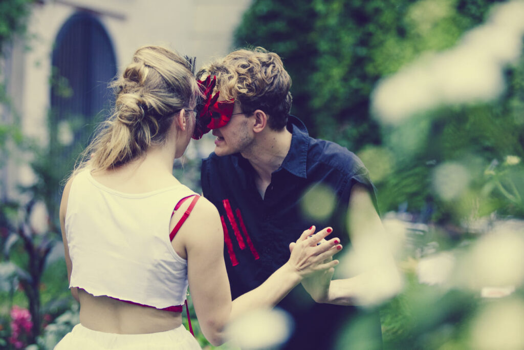 Auf dem Foto ist ein schwarz gekleideter Mann, der seine Hände mit der weiß gekleideten Frau verschränkt. Sie beide tragen rote Masken.