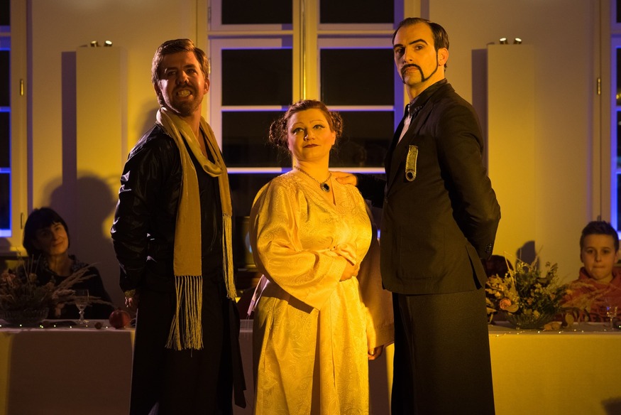 Auf dem Foto steht eine hell gekleidete Frau zwischen zwei schwarz gekleideten Männern mit stolzer Haltung vor einem Bankett.