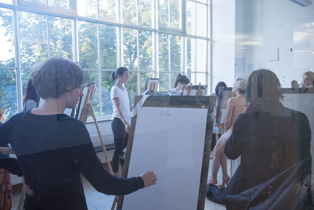 Auf dem Foto sind mehrere Menschen vor einem großen Fenster, die auf Staffeleien eine Person skizzieren.