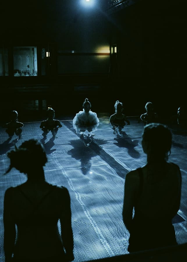 Auf dem Foto sind die Silhouetten des Publikums im Wasser zu sehen. Zwischen ihnen ist eine Performerin in weißem Tüllkleid.
