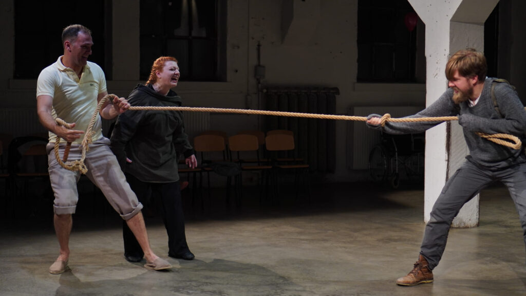 Auf dem Foto sind zwei Männer zu sehen die an den zwei Enden eines Stricks ziehen. Auf einer Seite steht eine schwarz gekleidete Frau die zuruft.