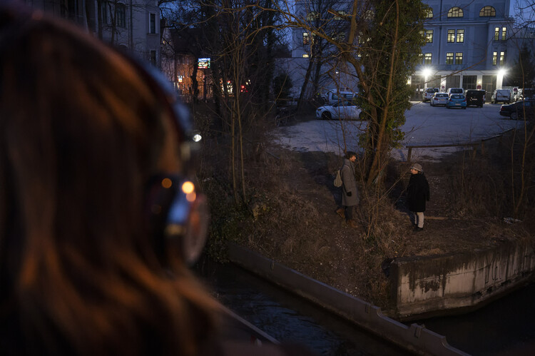 Auf dem Foto blickt man am Hinterkopf einer Person mit Kopfhörern vorbei und sieht zwei Menschen auf einem Stück Natur am Wasser stehen.
