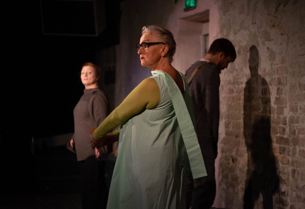 Auf dem Foto ist eine grün gekleidete Frau mit schwarzer Brille zu sehen. Hinter ihr ist ein Mann, der gegenüber einer Wand steht.