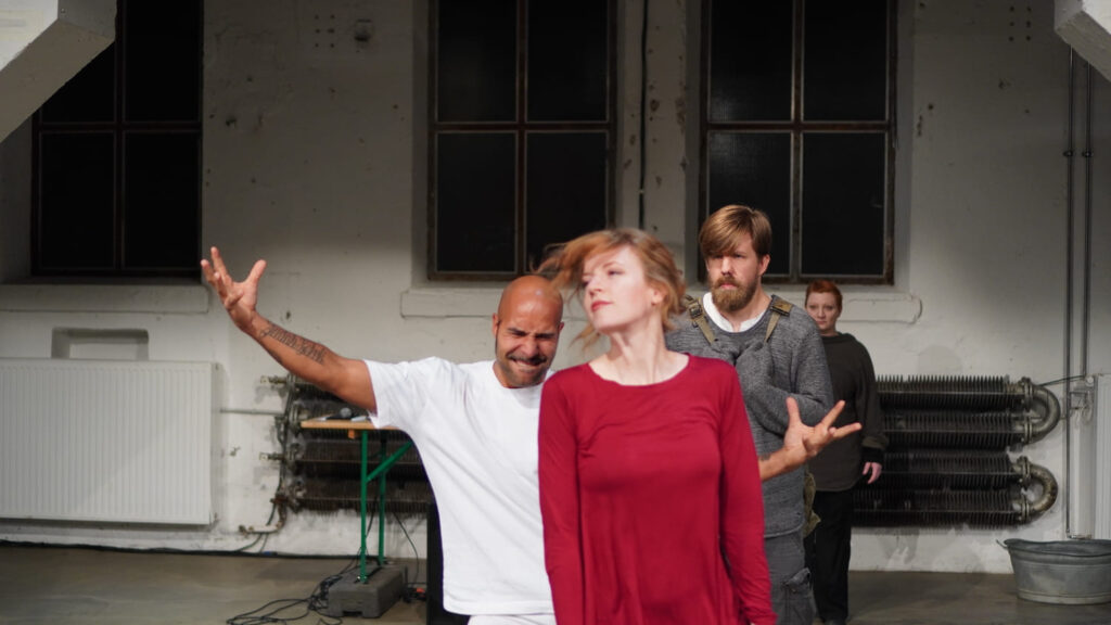 Auf dem Foto ist eine rot gekleidete Frau zu sehen. Hinter ihr steht ein weiß gekleideter Mann mit geöffneten Armen und einem angestrengtem Gesichtsausdruck. Ein grau gekleideter Mann in zusammengezogener Haltung steht hinter ihnen.