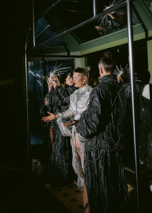 Auf dem Foto sind 2 Menschen in schwarzen Kitteln zu sehen. Zwischen ihnen steht jemand in silbrigem Kleid und offenen Armen.