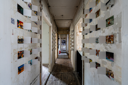 Auf dem Foto ist ein Hausflur zu sehen. An den weißen Wänden links und rechts sind viele aufklappbare, kleine Bilder.