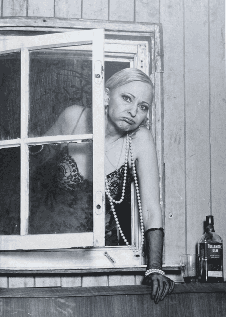 Auf dem schwarzweißen Foto ist eine Frau in Negligé und Perlenschmuck, die hinter einem offenen Fenster hervorsieht.