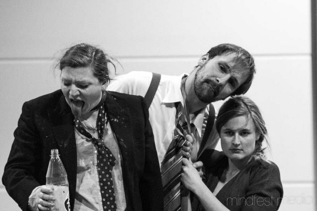 Auf dem schwarzweißen Foto ist eine Frau in Anzug und Flasche in der Hand, angeekelt ihre Zunge ausstreckt. Neben ihr ist eine Frau, die an der Krawatte eines Mannes zieht.