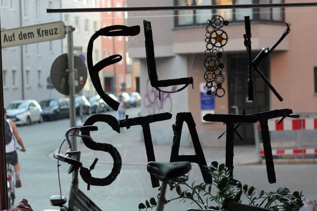 Auf dem Foto blickt man durch ein Schaufenster auf die Straße. Davor hängt in verschiedenen Fahrradteilen ein Schriftzug "FLIKSTATT"