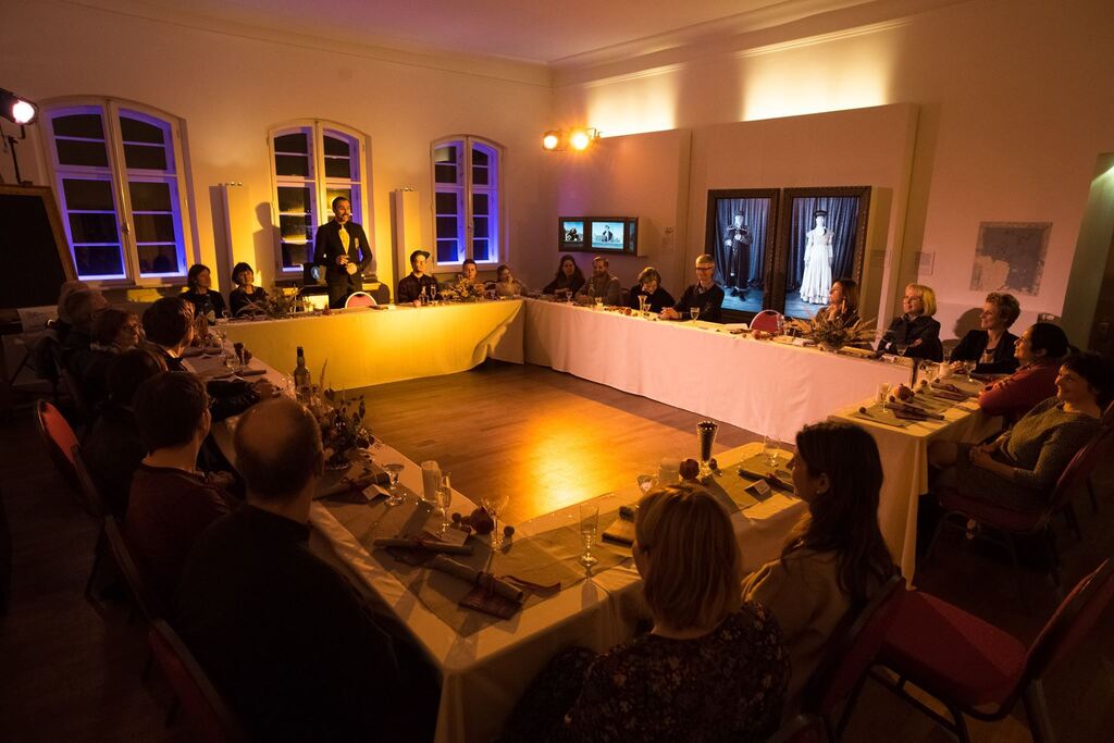 Auf dem Foto sitzen mehrere Menschen an einer o-förmigen Tafel und blicken zu einem Mann in schwarzem Anzug.
