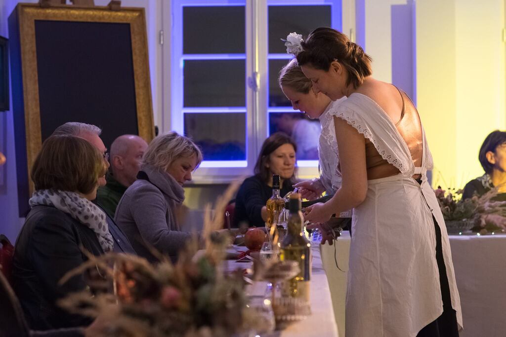 Auf dem Foto sitzt das Publikum an einem langen Tisch und wird von zwei Frauen in weißen Spitzenschürzen bedient.