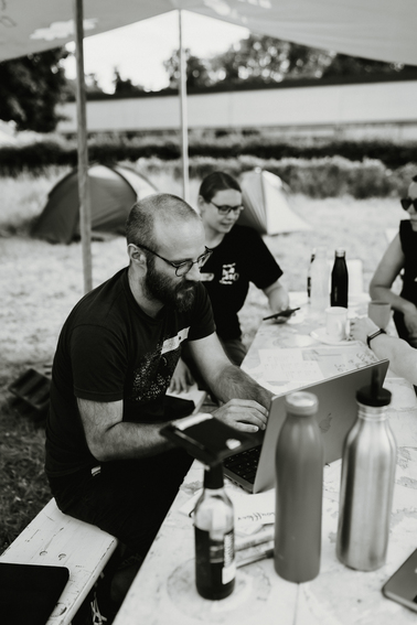 Auf dem schwarzweißen Foto sieht man einen Mann draußen vor einem Computer sitzen. Hinter ihm sitzen zwei sich unterhaltende Frauen und zwei Zelte im Rasen.