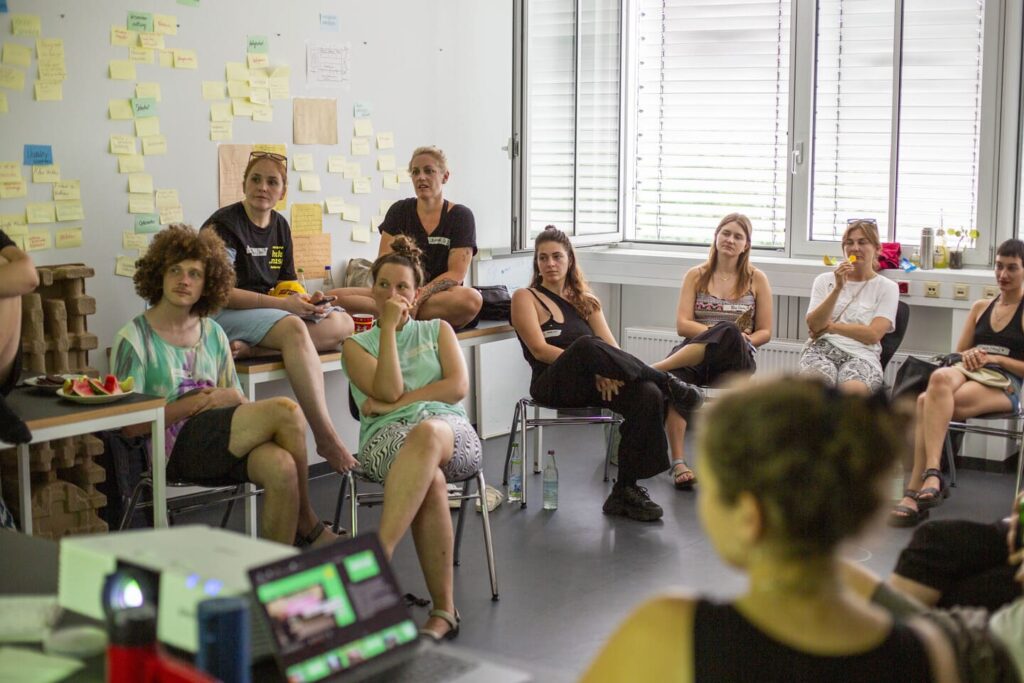 Auf dem Foto sieht man eine Gruppe auf Tischen und Stühlen sitzen. Sie blicken konzentriert in eine Richtung und hinter ihnen ist eine Wand mit vielen Post-its.