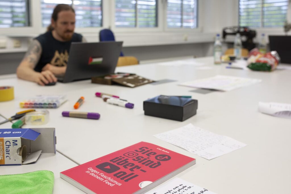Auf dem Bild sieht man einen Mann an einem großen Tisch vor einem Computer. Vor ihm liegt ein rotes Buch mit dem Titel "Sie sind überall – Gegen Faschismus in deinem Feed".