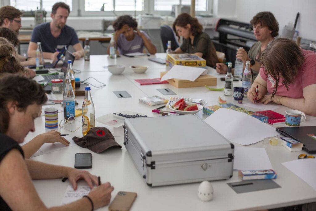 Auf dem Foto ist eine Gruppe an einem bunt beladenen Tisch. Sie sind konzentriert am Schreiben.