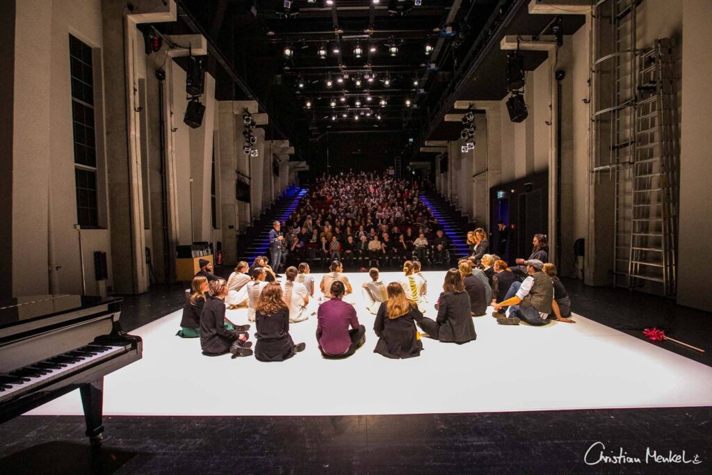 Auf dem Foto sitzen mehrere Menschen auf einer weißen Fläche. Sie blicken zum Publikum hoch.