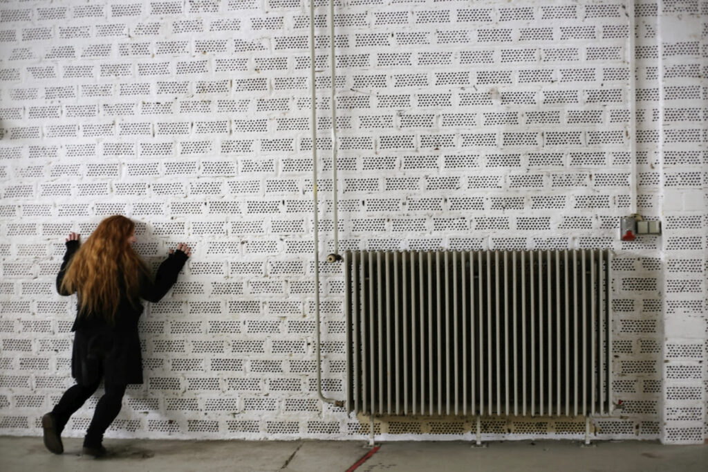 Auf dem Foto ist eine Heizung an einer Backsteinwand. Links davon steht eine schwarz gekleidete Frau mit ihren Händen an der Wand.