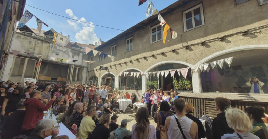 Auf dem Foto sind viele Menschen in einem Innenhof versammelt. Über ihnen hängen bunte Wimpel und vor ihnen ist eine kleine Bühne.