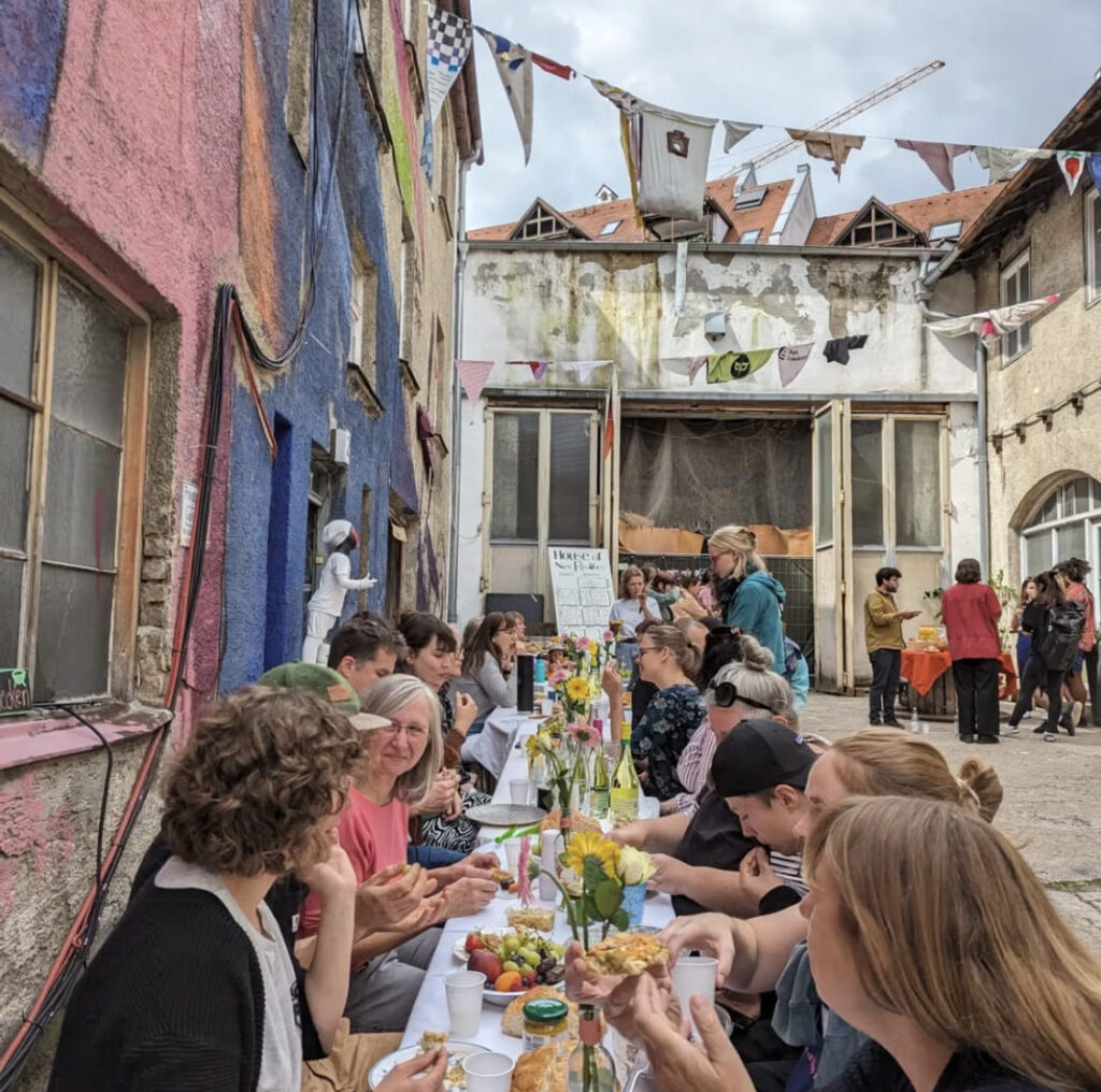 Auf dem Foto sieht man einen langen Tisch an einer bunten Hauswand. Viele Menschen sitzen dort und genießen ihr Essen.