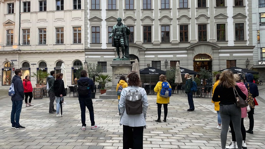 Auf dem Foto stehen mehrere Menschen auf einem Platz um eine Statue. Die Statue stellt einen edel gekleideten Mann in stolzer Haltung dar.