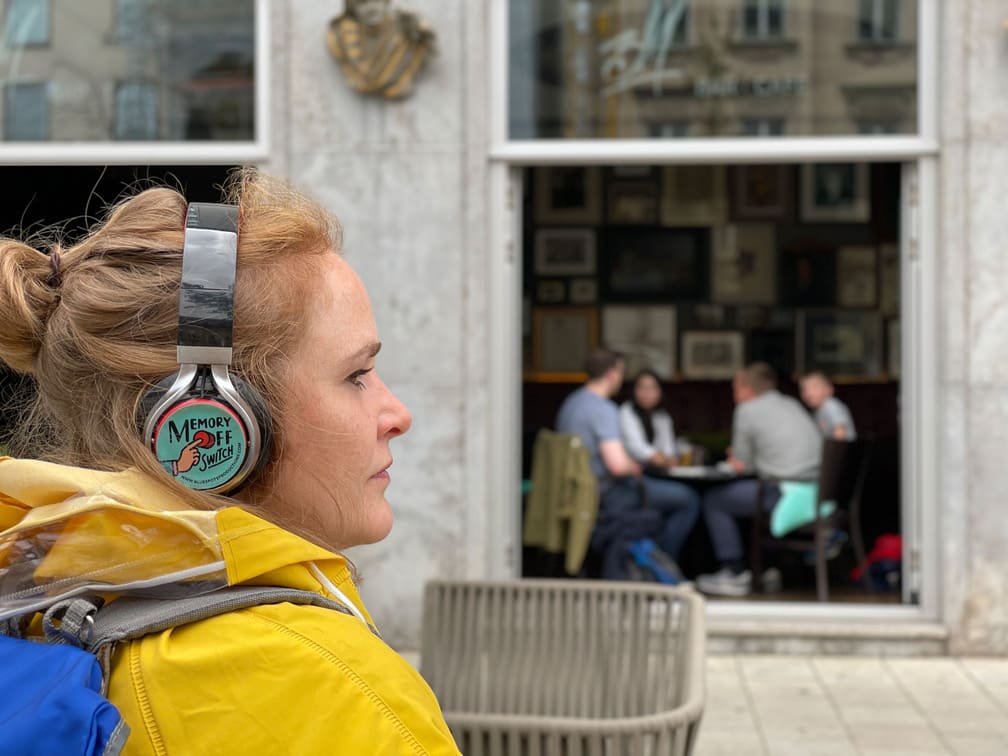 Auf dem Foto ist eine Frau mit gelber Jacke vor einem Café. Sie trägt Kopfhörer mit der Aufschrift "MEMORY OFF SWITCH"