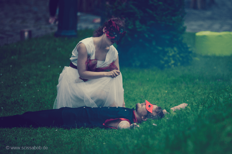 Auf dem Foto ist ein schwarz gekleideter Mann mit roter Maske liegend in grünem Rasen. Neben ihm kniet eine Frau in roter Maske und weißem Kleid.