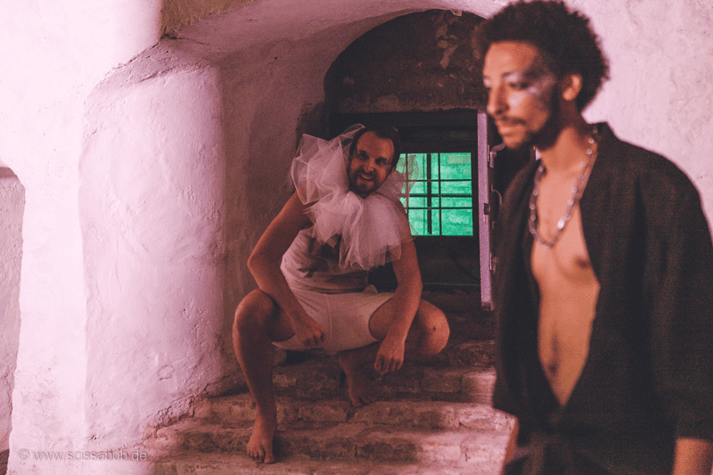 Auf dem Foto ist ein Mann mit weißer Halskrause in der Hocke auf einer kleinen Treppe. Vor ihm steht ein schwarz gekleideter Mann mit nachdenklichem Blick.