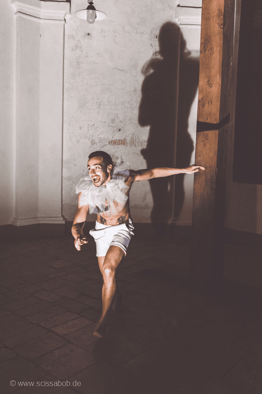 Auf dem Foto ist ein aufgeregter, schreiender Mann in weißer Hose und Halskrause. Hinter ihm ist sein großer Schatten an der Wand.