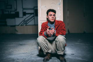 Auf dem Foto sitzt ein Mann an einer Säule und blickt in die Kamera. Er trägt eine rote Jacke und scheint etwas in den Händen zu haben.