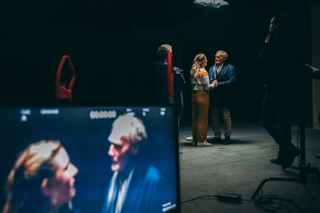 Auf dem Foto ist die Szene von zwei Menschen, die sich an den Händen halten. Im Vordergrund ist dieselbe Szene auf einem Bildschirm zu sehen.