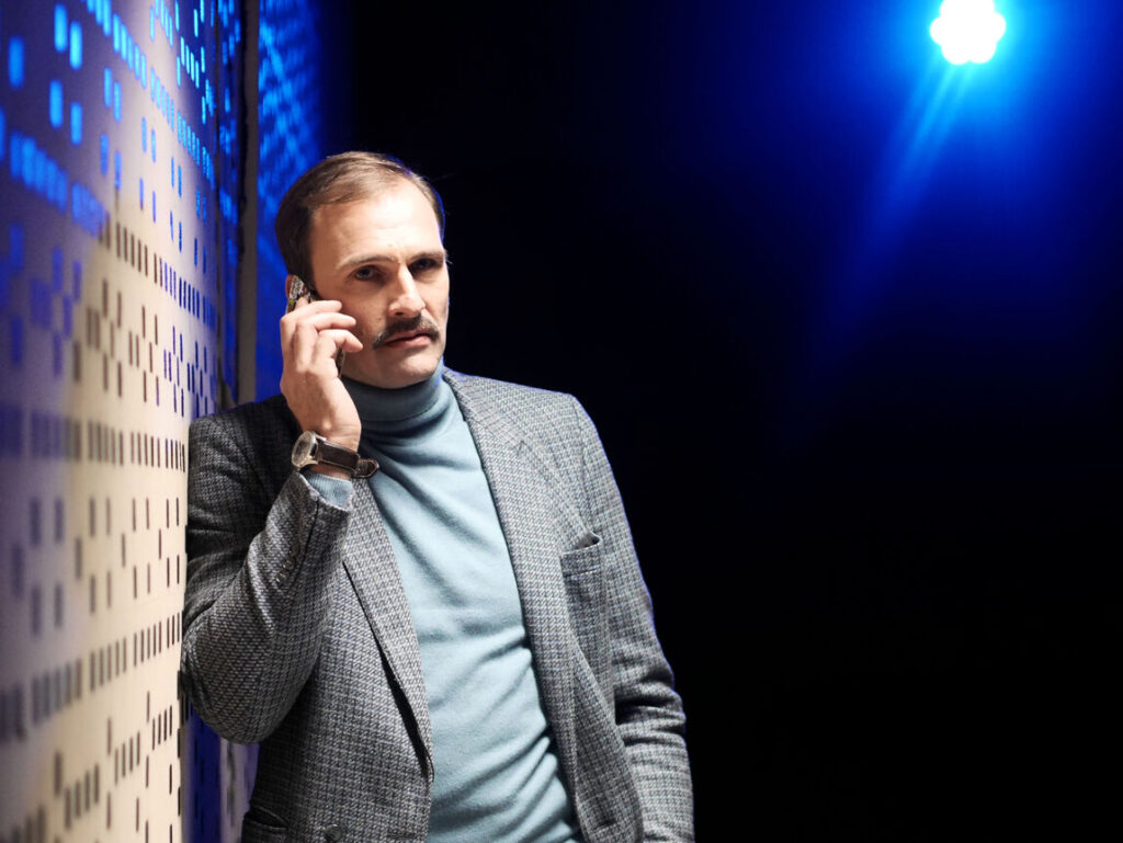 Auf dem Foto ist ein telefonierender Mann mit kariertem Sakko und Rollkragen. Er lehnt an einer gemusterten Wand, die von einem blauen Licht angestrahlt wird.