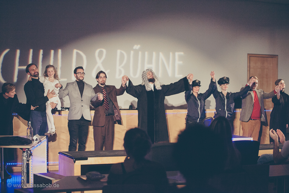 Auf dem Foto sind mehrere kostümierte Menschen die ihre Hände dankend in die Luft strecken. Hinter ihnen ist der Schriftzug "SCHULD UND BÜHNE".