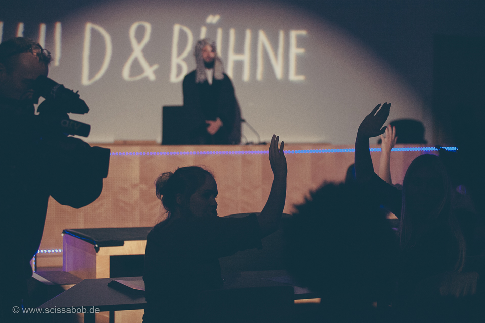 Auf dem Foto ist die Silhouette von einem Publikum mit Meldungen. Hinter ihnen steht ein Mann mit weiß-lockiger Perücke, hinter dem der Schriftzug "SCHULD UND BÜHNE" zu sehen ist.