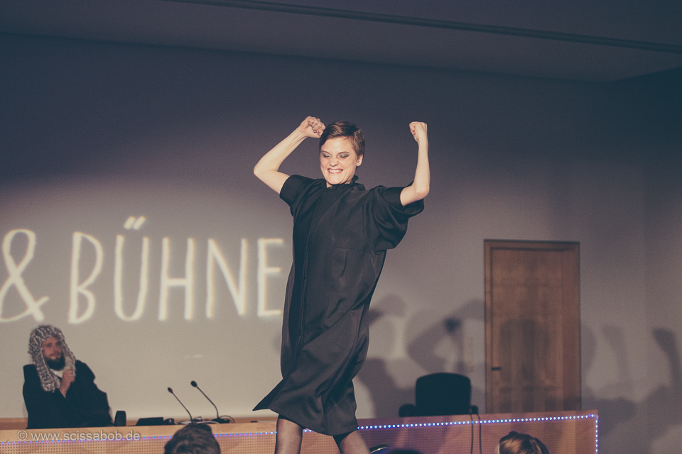 Auf dem Foto ist eine schwarz gekleidete Frau die ihre Arme mit Fäusten in die Luft streckt. Sie hat ein glückliches Gesicht und hinter ihr ist der Schriftzug "SCHULD UND BÜHNE".