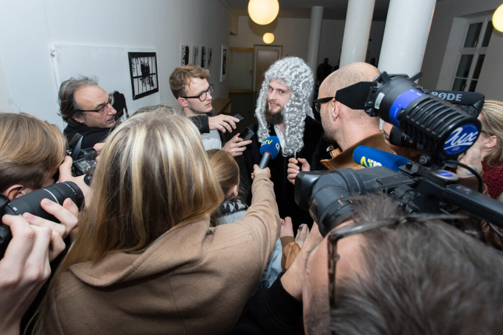 Auf dem Foto ist ein Mann mit weiß-lockiger Perücke, der inmitten einer Gruppe von Reporter:innen mit Mikrofonen und Kameras steht.