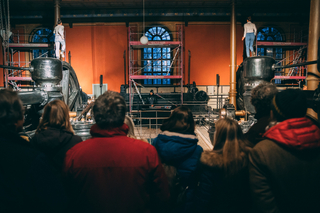 Auf dem Foto ist eine Bühne mit Baustellenkonstruktionen. Darauf stehen zwei Menschen. Vor der Bühne sieht man die Rücken des Publikums.